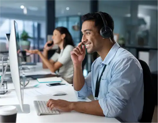 Male technical support representative with a headset smiling while assisting a client, seated at a desk in a modern office.