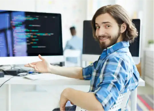 Young male software developer in a blue plaid shirt sitting at a desk with multiple monitors displaying code, smiling and gesturing towards the screen.