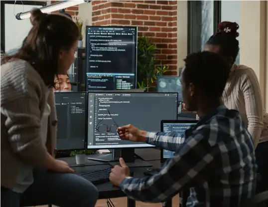 Group of diverse software developers collaborating at a desk with multiple monitors displaying code, discussing a project.
