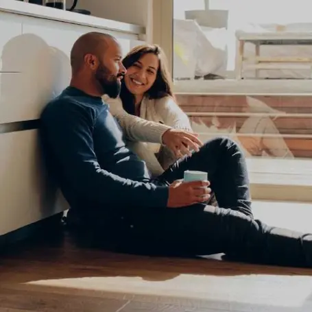 Man wearing blue shirt and dark pants sitting on the floor next to a woman in a white shirt, leaning against white cabinetry.