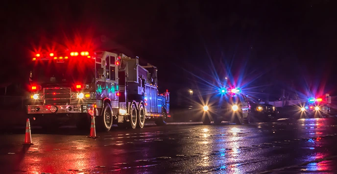 Firetruck with emergency lights on, behind two orange traffic cones. Three police SUVs behind the firetruck, all with lights on. Late at night on a highway.